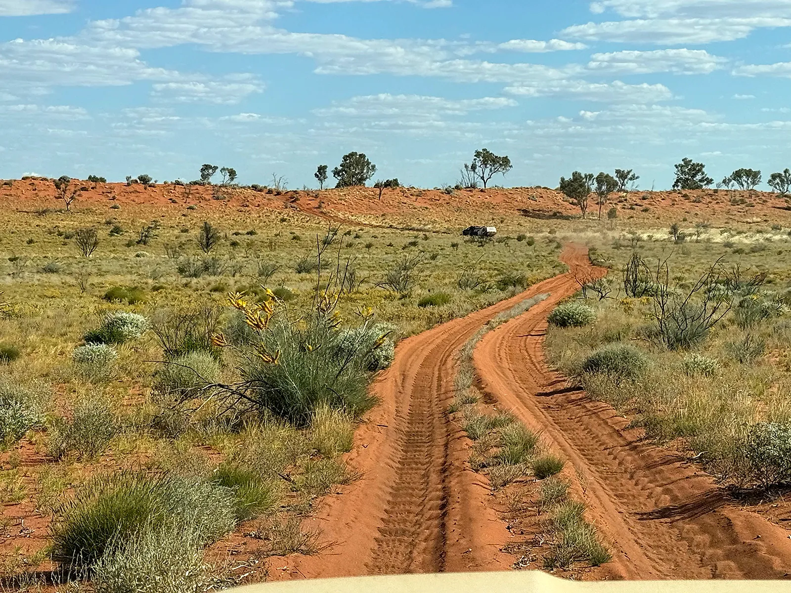 Deep wheel ruts in a red sandy track through low scrub, with a four-wheel drive and camper in the distance