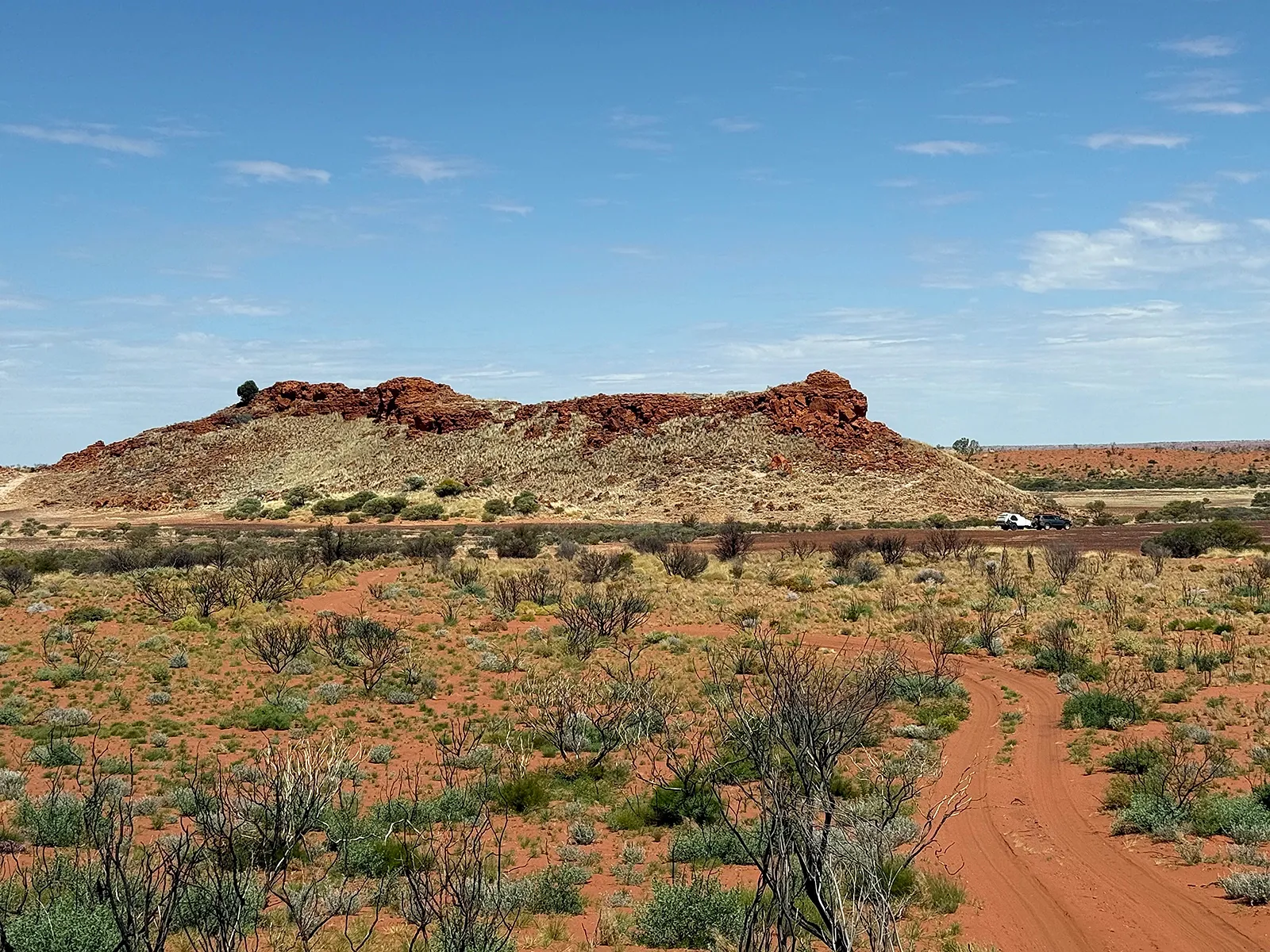 Red dirt track winding through desert scrub past a rocky red outcrop, with two four-wheel drives in the distance