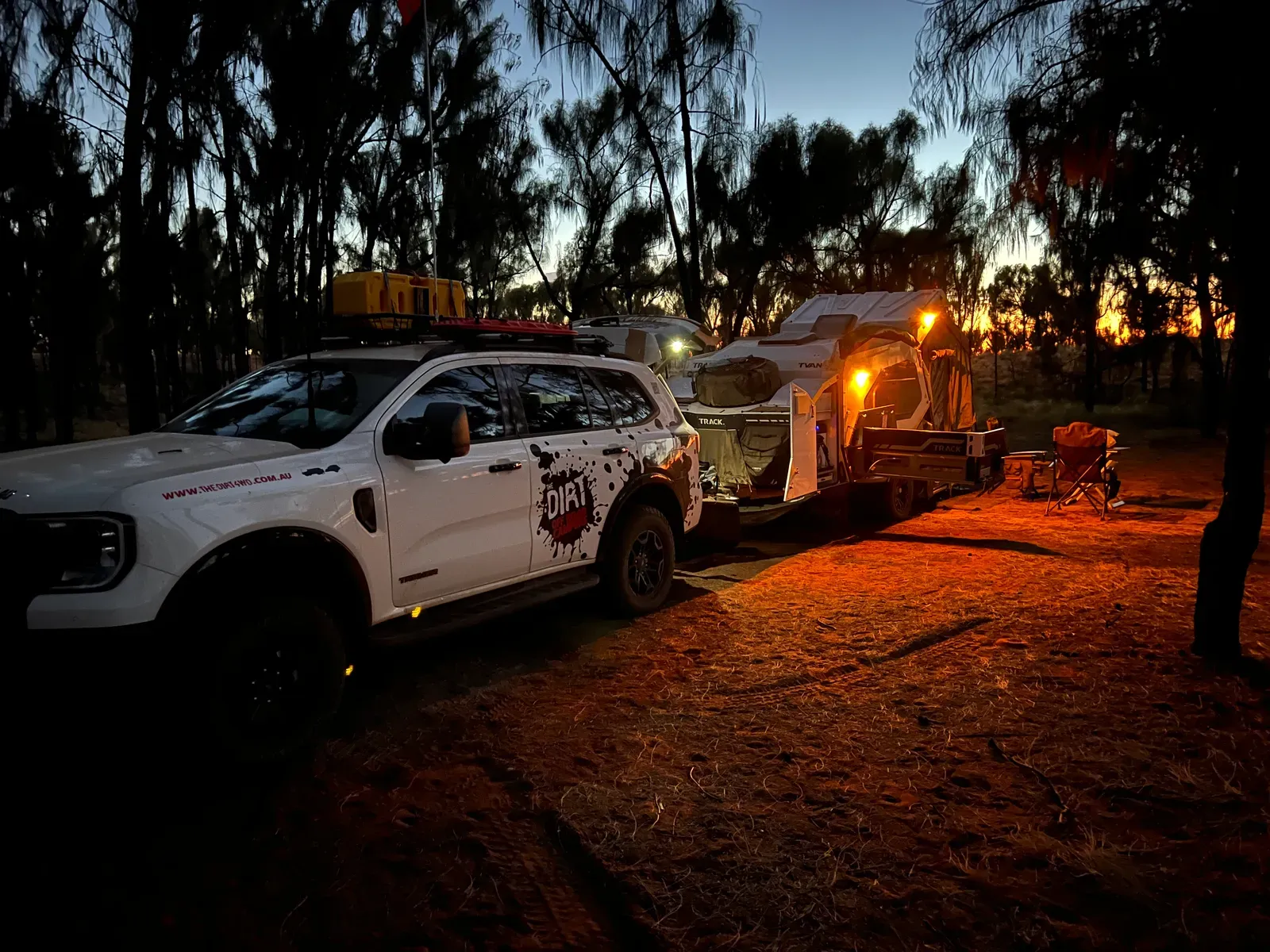 A Ford Everest towing a Track Trailer Tvan parked beneath trees at dusk, with the camper lights glowing