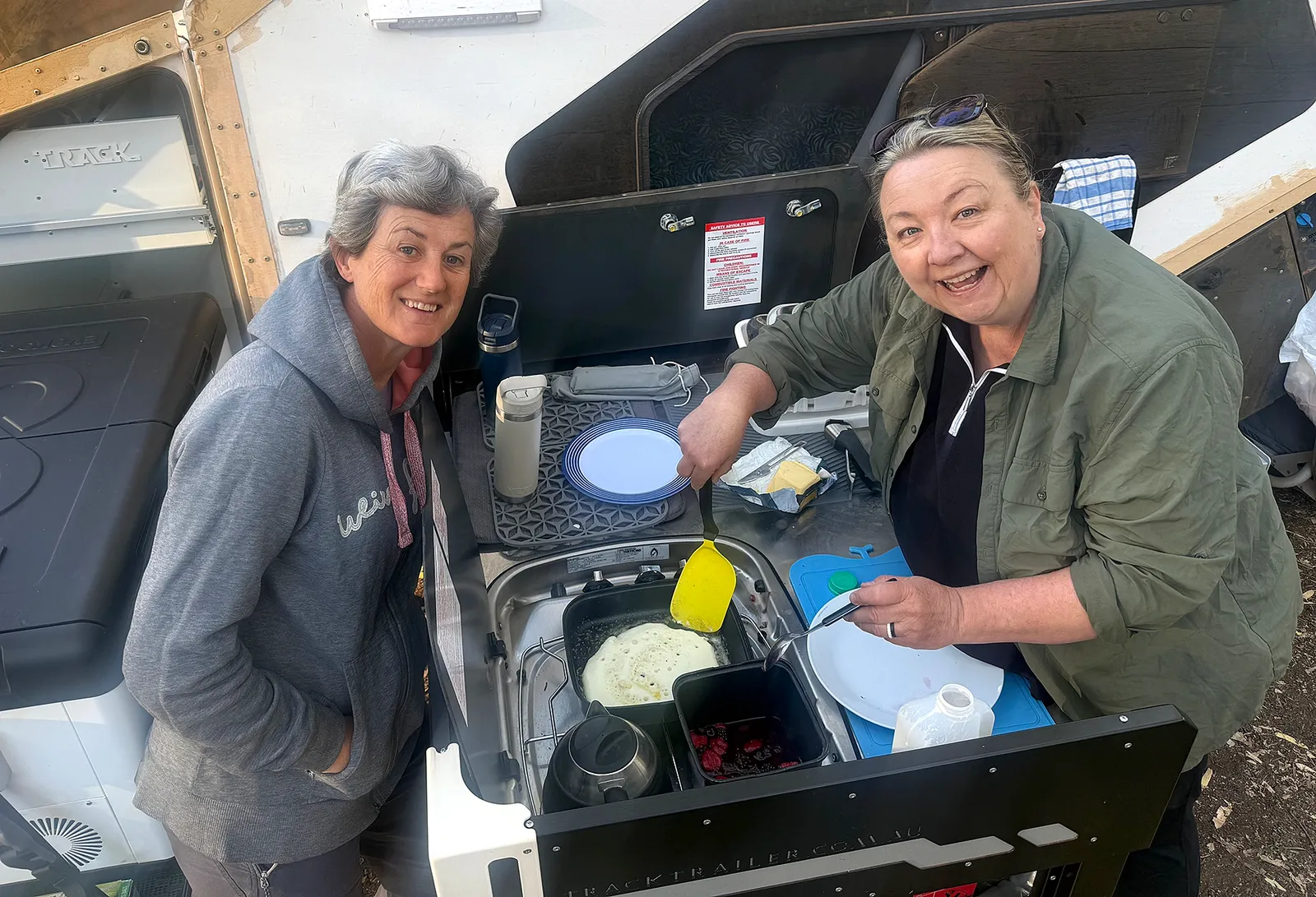 Two women preparing food at the open kitchen of a Track Trailer Tvan, with one stirring a pan